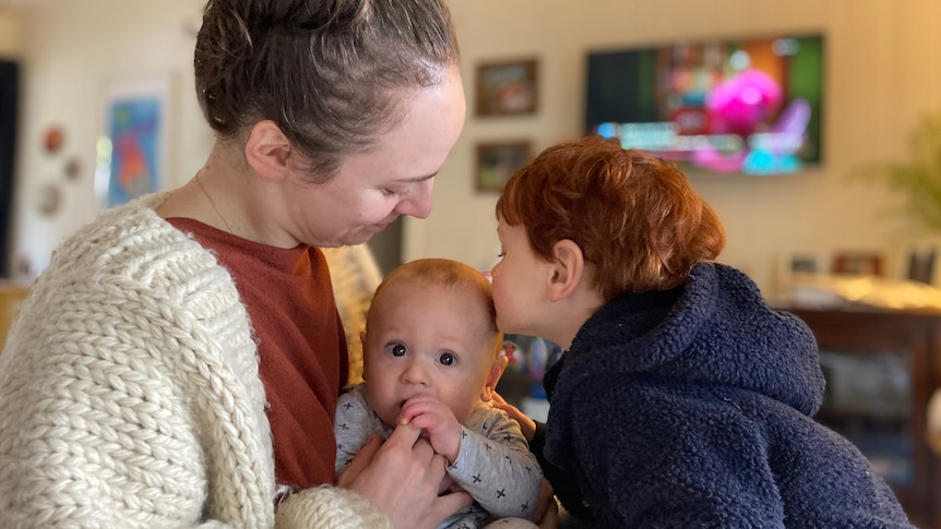 A woman sits with her baby in her living room as her young son gives the baby a kiss.