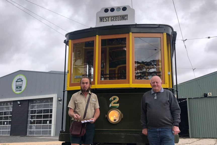 Two men smile in front of a green and yellow tram