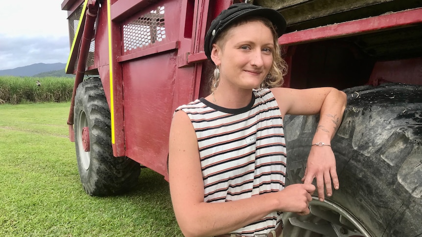 A young woman with short blonde hair, a tank top and a black hat leans on the wheel of a power-haul vehicle.