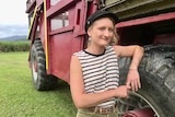 A young woman with short blonde hair, a tank top and a black hat leans on the wheel of a power-haul vehicle.