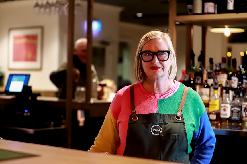 Woman wearing bright coloured jumper and apron stands at bartop with pub in background