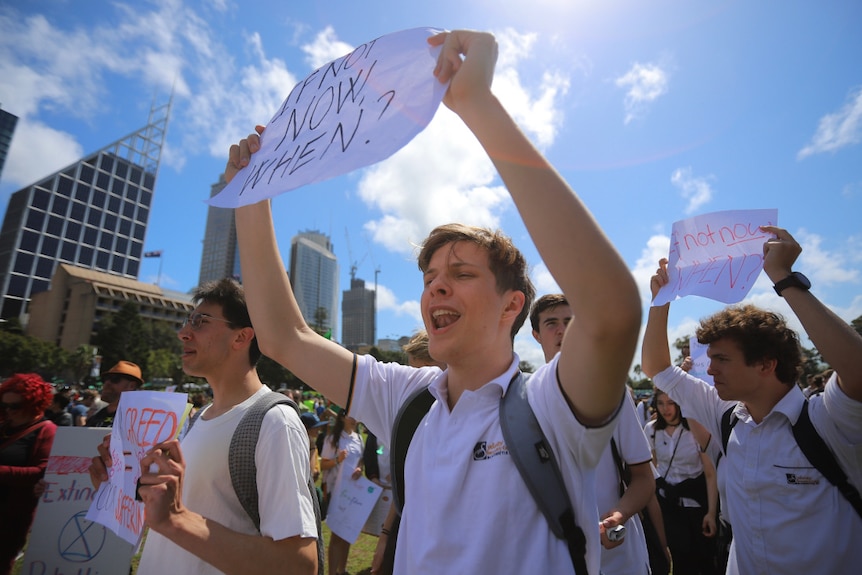 A boy with a white polo shirt and backpack holds up a sign that says  'if not now, when?'