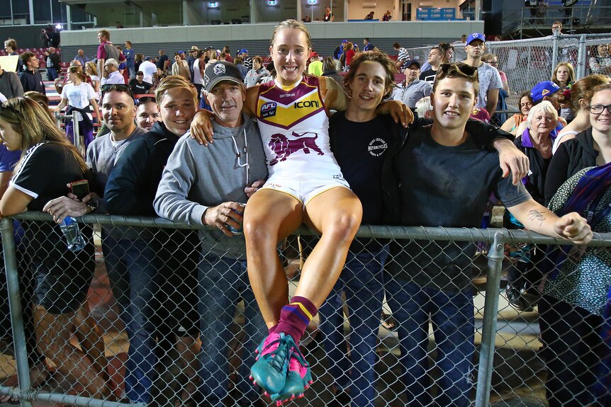 Jacqui Yorston sits on the fence and poses with supporters in a Brisbane Lions guernsey