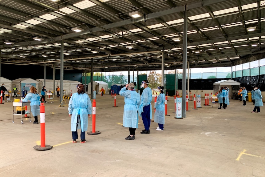 Healthcare workers in full PPE wait for cars to arrive at a drive-through vaccination site.