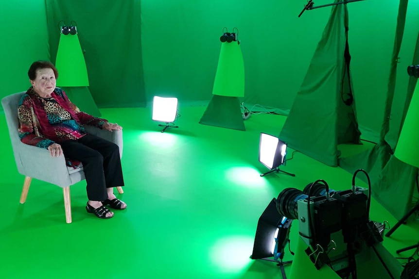 An old woman with brown hair sits in a room surrounded by green screens and cameras.