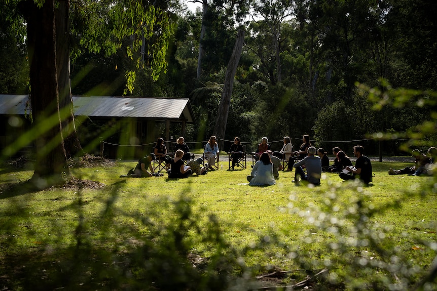 A meeting takes place in amongst gum trees and green grass on a sunny day at a site within the former reserve of Coranderrk