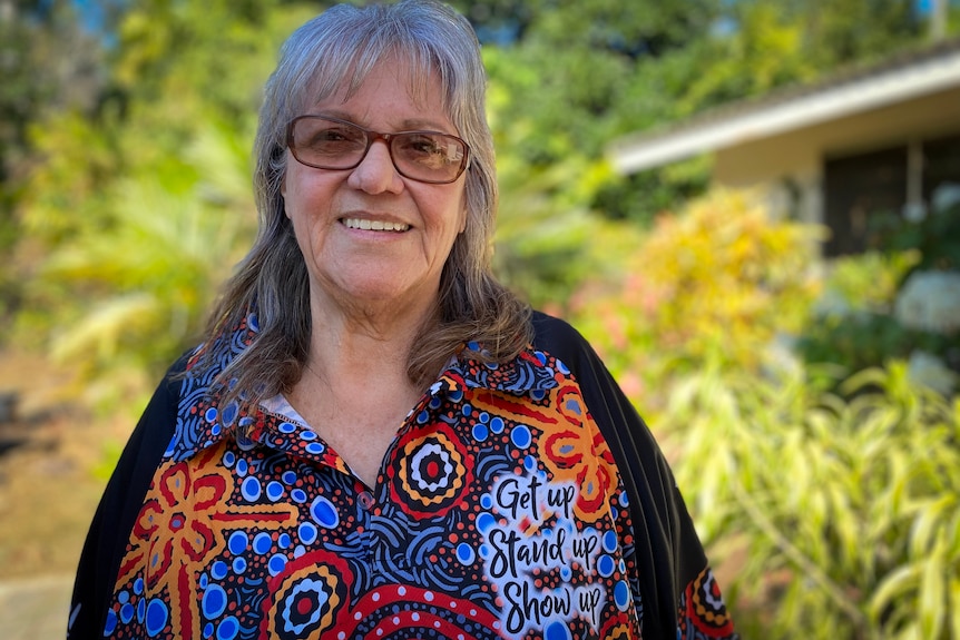 A portrait of a woman in her garden wearing a NAIDOC week shirt.