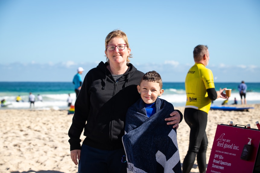 A boy holding a towel by the beach and his mother next to him with her arm around his shoulder.