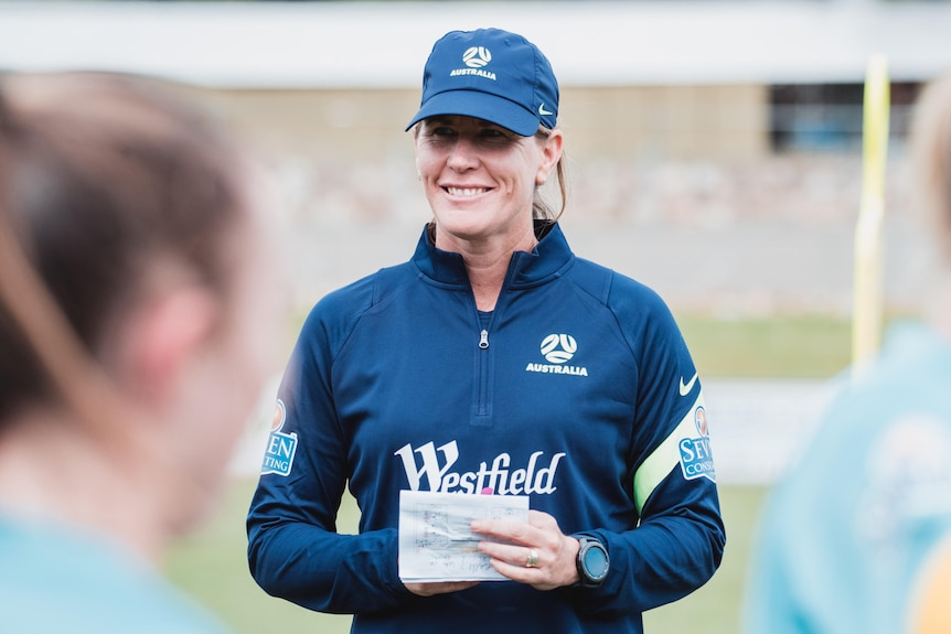 Mel Andreatta holds a piece of paper and smiles as she addresses players.