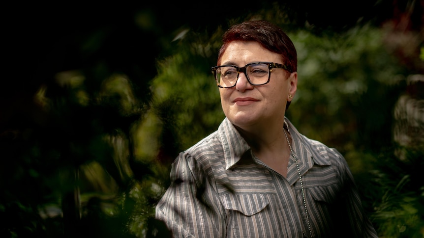 A middle-aged woman with short hair and glasses looking pensively by some bushes