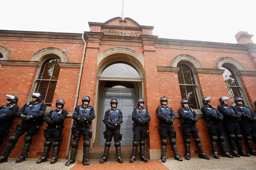 Victoria Police offices stand in front of a building wearing vests, glasses and helmets.
