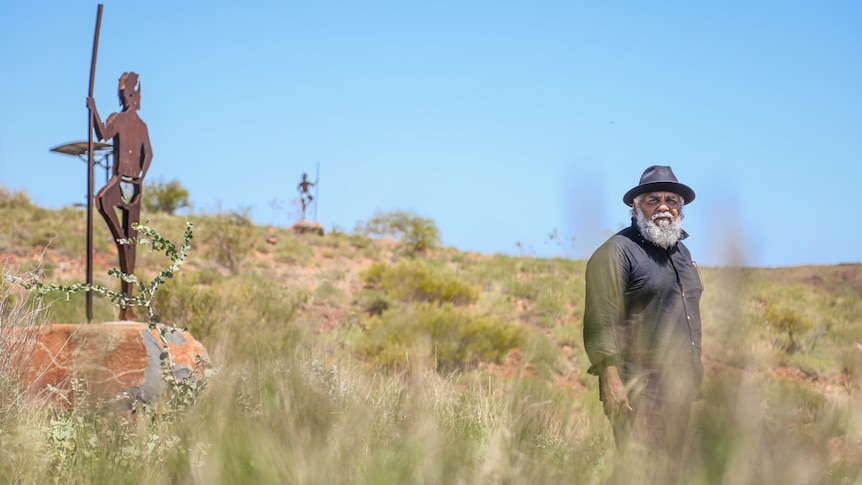 Long shot of an older indigenous man standing in tall grass near a statue.