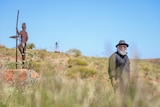 Long shot of an older indigenous man standing in tall grass near a statue.