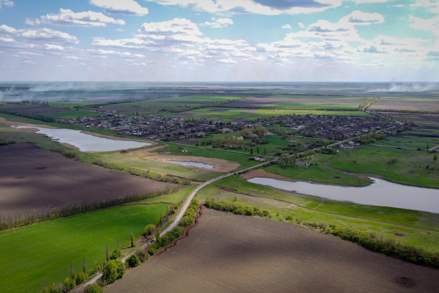 A bird's-eye view of a tiny village surrounded by green grass and a large lake.