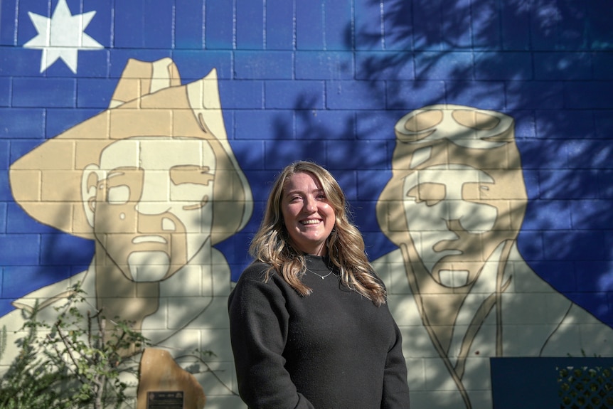 A woman with blonde, wavy hair smiles in the sunlight against a mural commemorating veterans.