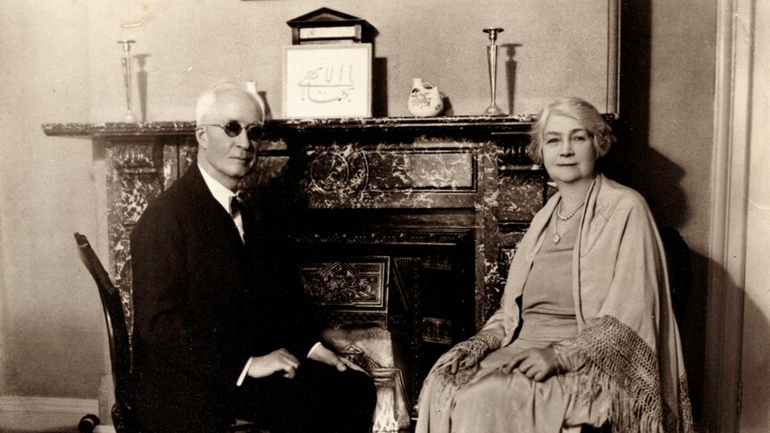 Black and white photo from early 20th century of man and woman dressed formally and sitting in front of fireplace.