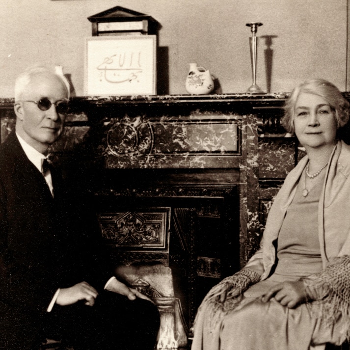 Black and white photo from early 20th century of man and woman dressed formally and sitting in front of fireplace.