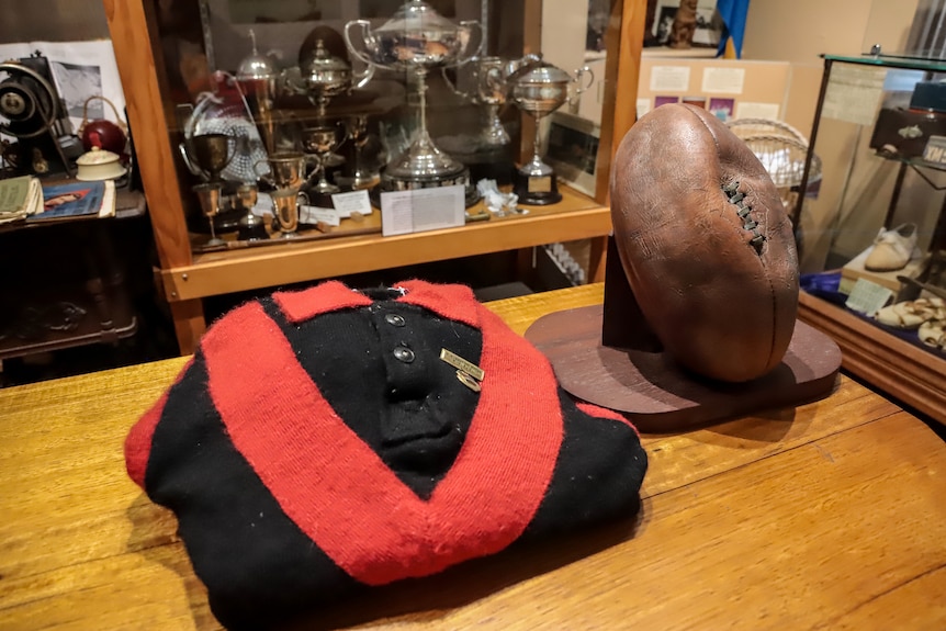 An old red v-striped black jumper and old leather ball sit on a wooden museum bench in front of trophy cabinet