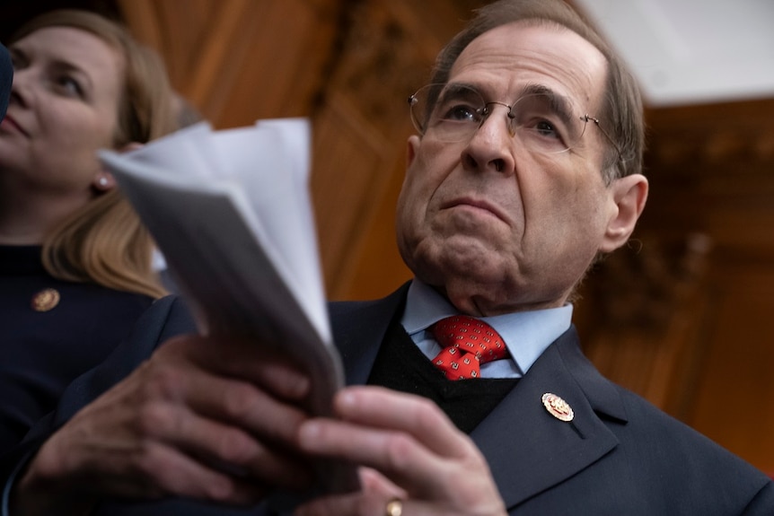 A close up image shows a man in a suit holding papers in front of his chest while he stands next to a woman.