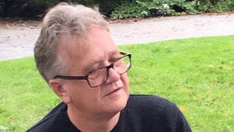 A bespectacled man sits on a bench with grass in the background.