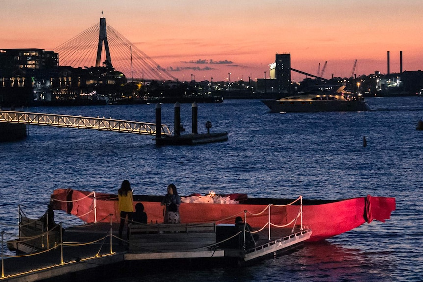 Large metal canoe moored at the end of a pontoon with Sydney Harbour in the background.