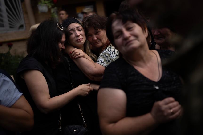 a crying woman is held by two other crying women as people gather to mourn the loss of her husband