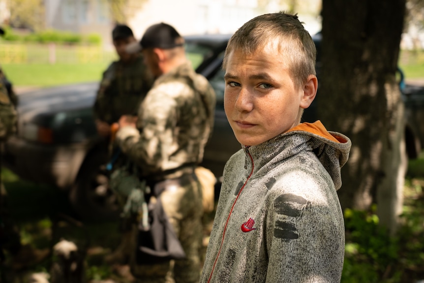 A boy wearing a jumper stares at the camera while surrounded by other people.