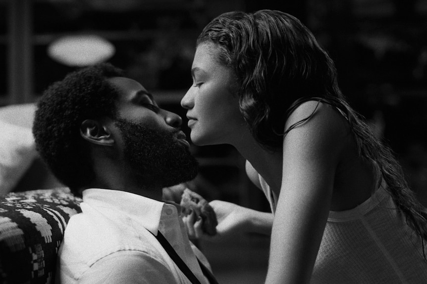 Black and white side-view of young man in white shirt on couch, straddled by young woman in white tank top, faces close.