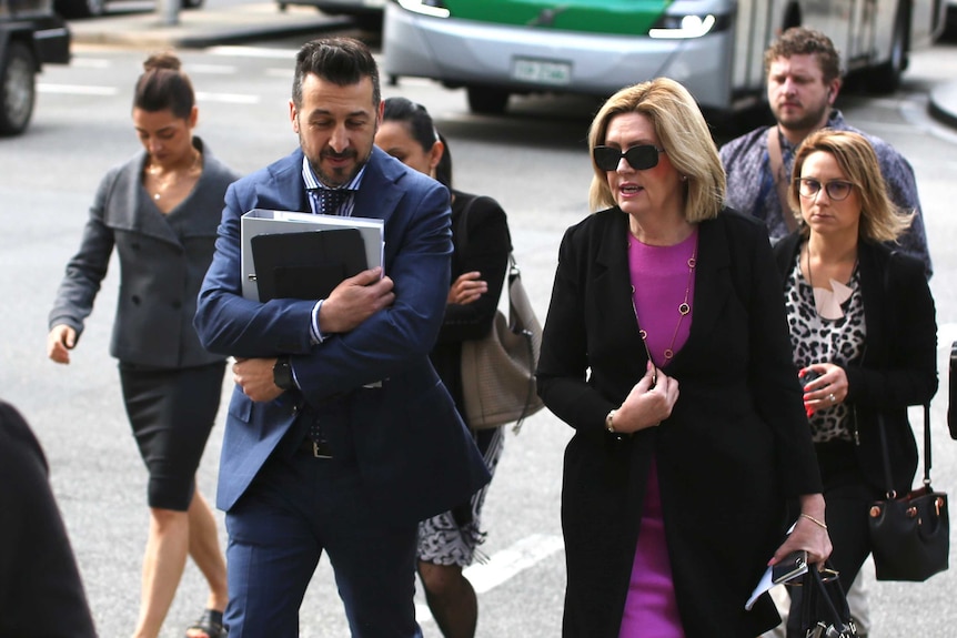 A group of people including Lisa Scaffidi walk along a busy sidewalk carrying bags and files of documents.