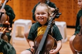 A primary school aged white girl in a green and yellow uniform smiles excitedly while seated holding a cello.