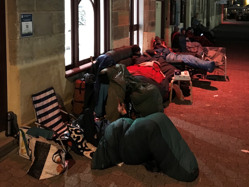 people in sleeping bags, camping chairs and couch in early hours of the morning sitting on pavement outside a gallery.
