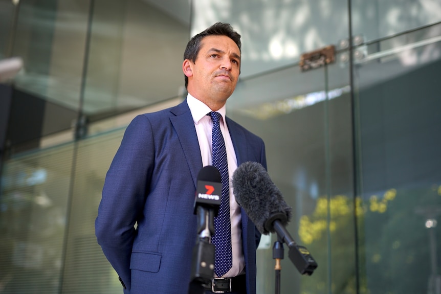 Blair Boyer standing with his hands behind his back in a navy suit with two microphones in front of him