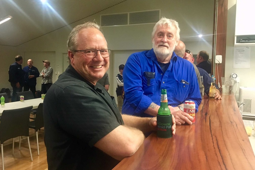 Two men lean against a bar drinking beer and smiling