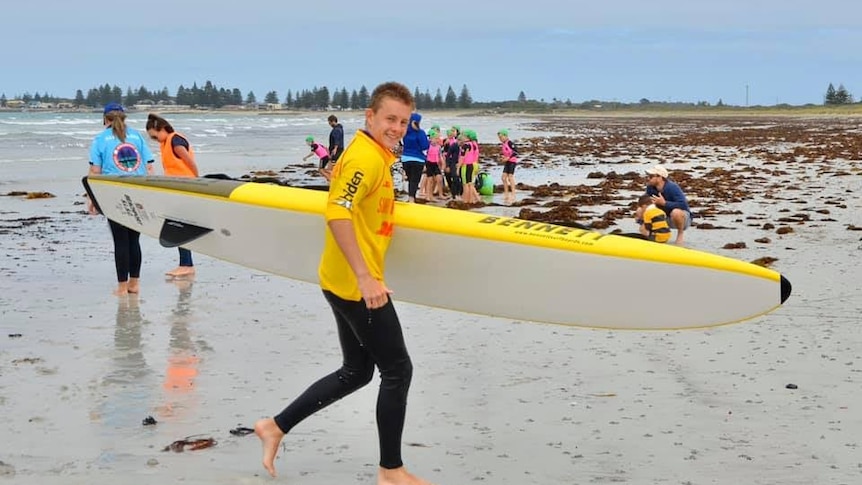 A teenage boy in a wetsuit and yellow swimming shirt runs across the sand carrying a board smiling.