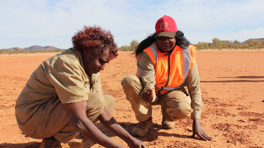 Two women crouch down in red dirt examining it.