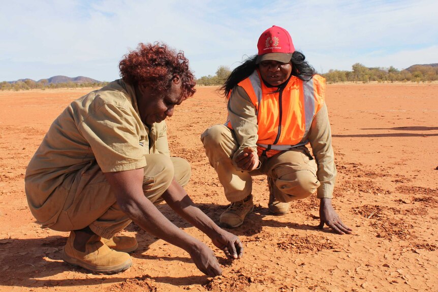 Two women crouch down in red dirt examining it.