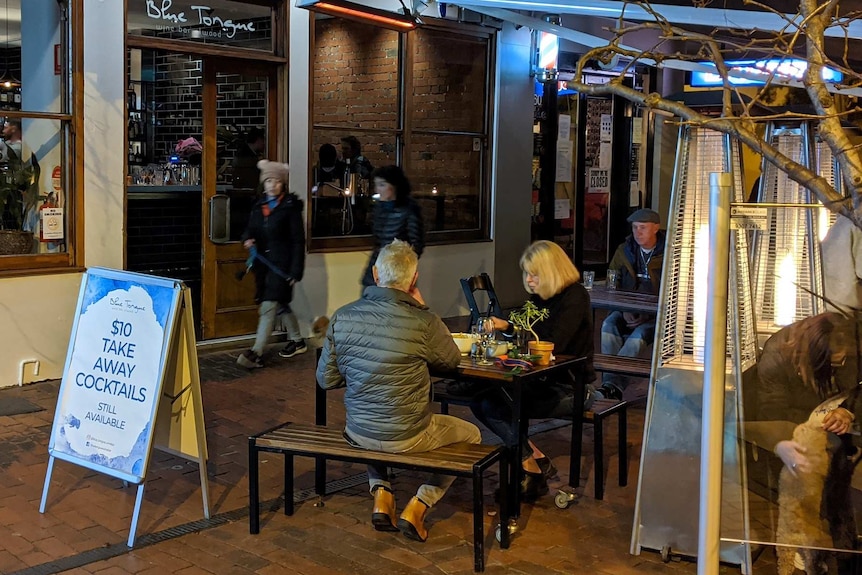 A couple sit outside near an outdoor heater having dinner under an awning.