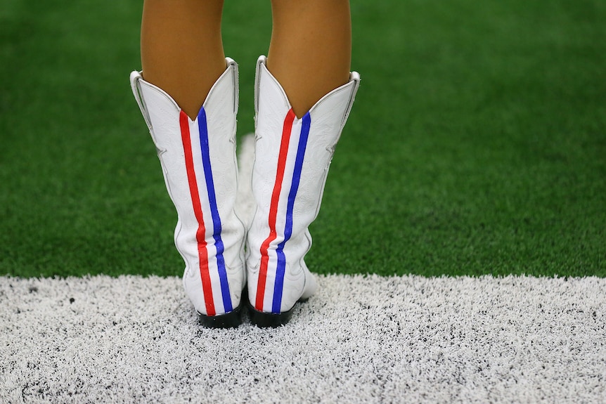 A Dallas Cowboys cheerleader's white booted feet stand on the sidelines at an NFL game.