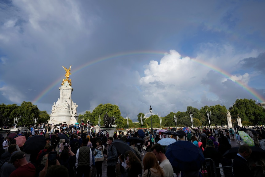 A crowd is gathered outside Buckingham Palace as two rainbows stretch across the sky. 