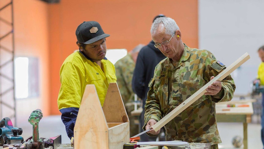 A man in army fatigues demonstrating some wood work to a young indigenous man