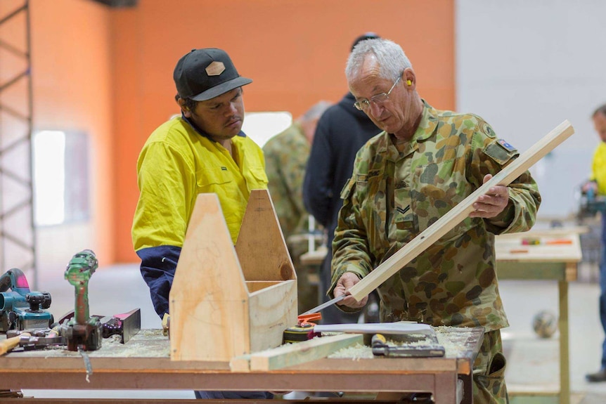 A man in army fatigues demonstrating some wood work to a young indigenous man