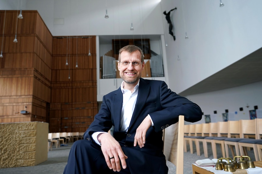 Jan Korditschke sits on a pew in a building with a crucifix on the wall. 