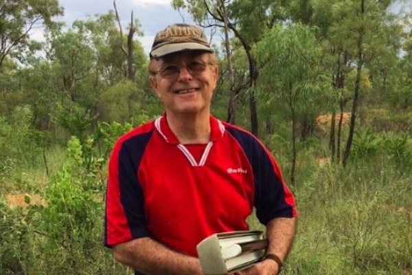 A smiling, bespectacled man wearing a cap stands in the bush holding a net.