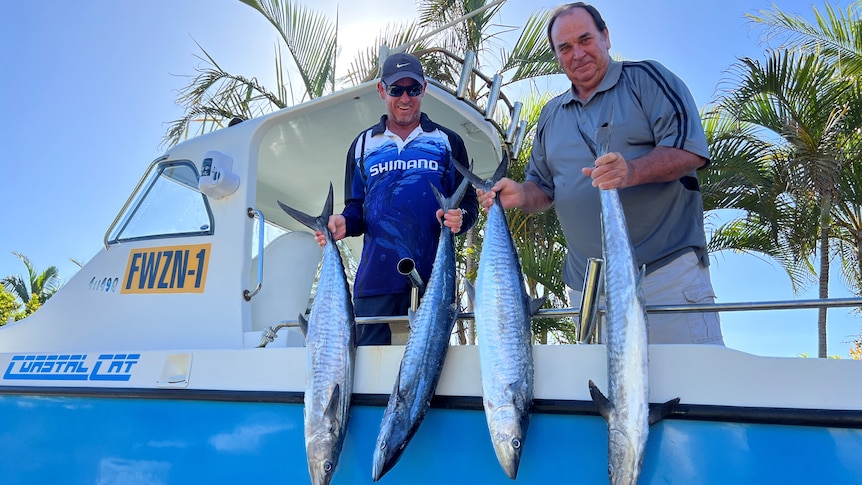 Two men dangling four large fish off the side of a blue boat on a blue-sky day and palm trees in the background