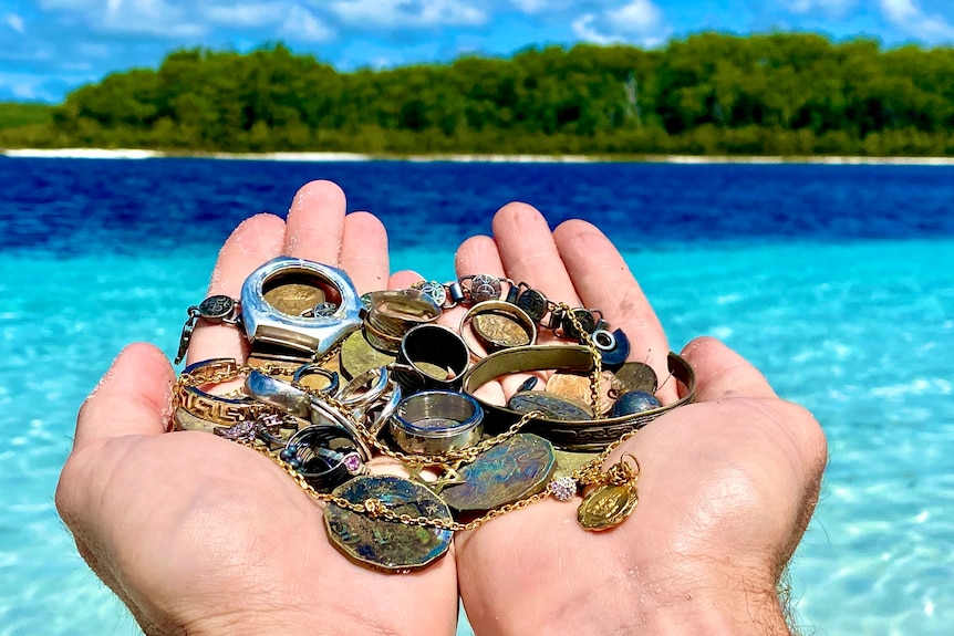 Man's hands holds a handful of rusted metal coins, bracelets, watch, ring, under a blue sky, green trees, beach in distance.