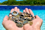 Man's hands holds a handful of rusted metal coins, bracelets, watch, ring, under a blue sky, green trees, beach in distance.