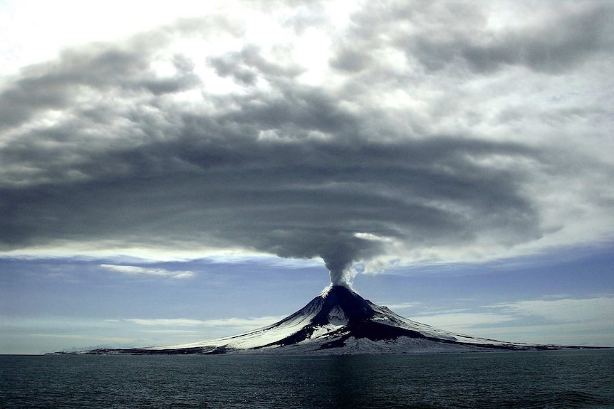 A volcano surrounded by ocean erupting with huge grey clouds of ash radiating above it