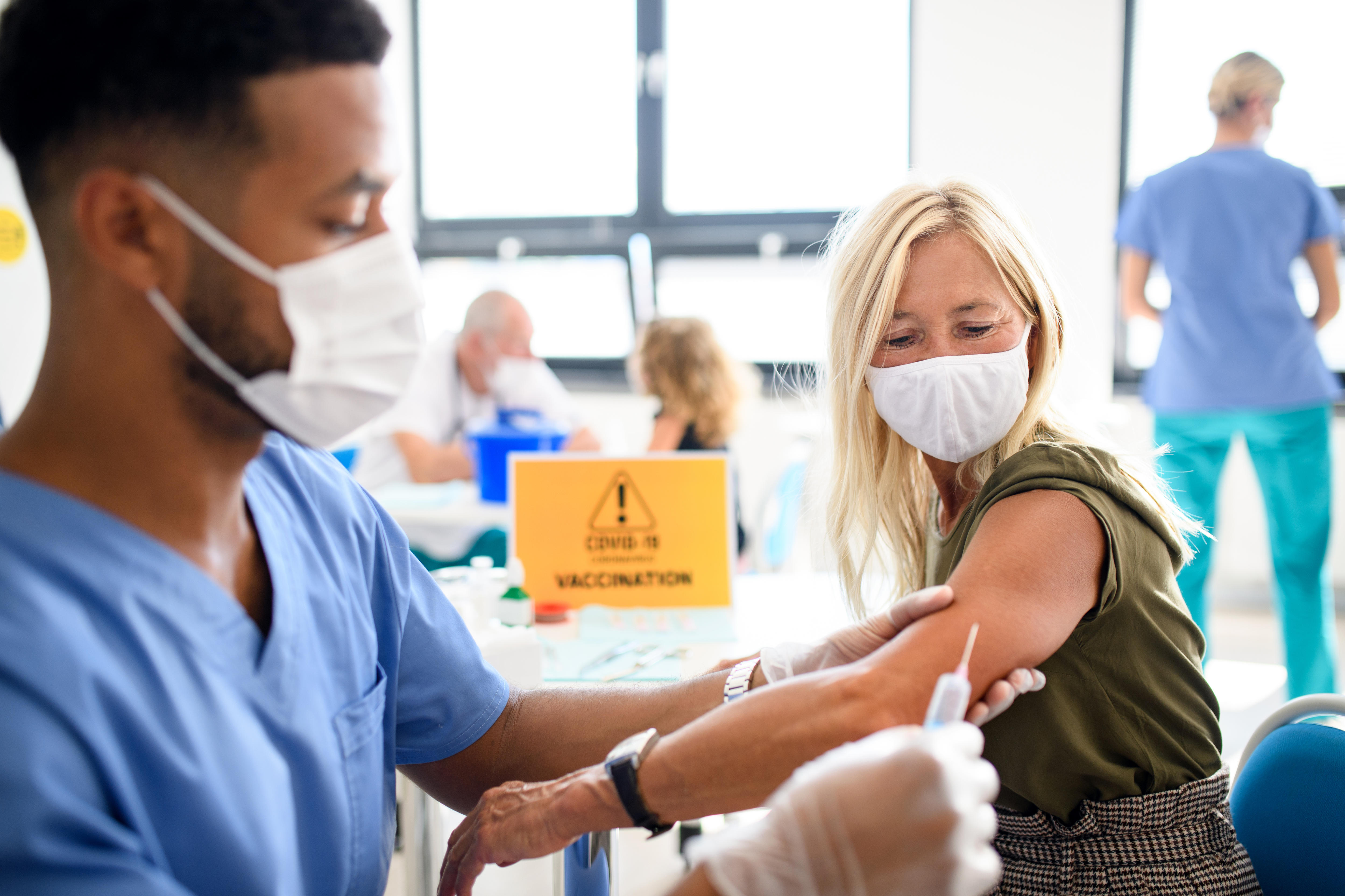 A man in blue scrubs and a white face mask is holding the arm of a woman in preparation for a vaccine.