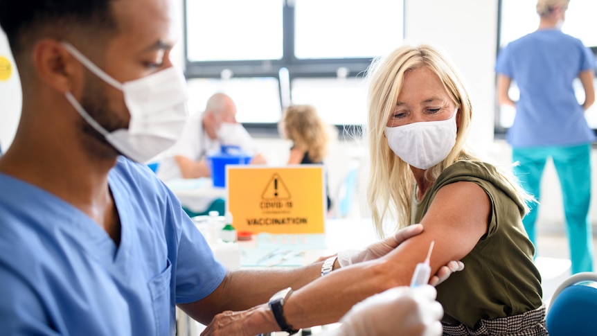 A man in blue scrubs and a white face mask is holding the arm of a woman in preparation for a vaccine.