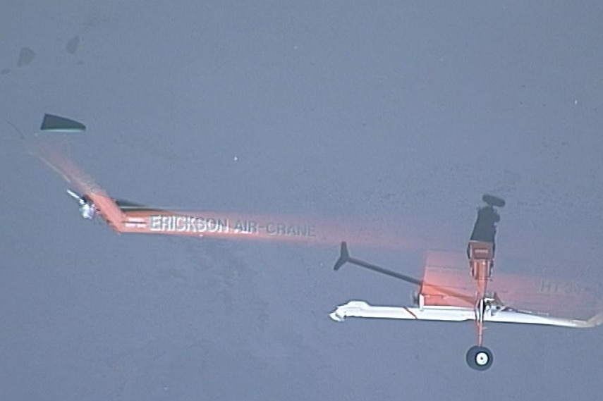 An aerial view of an orange helicopter lying mostly submerged in a body of water bordered by dense bushland.